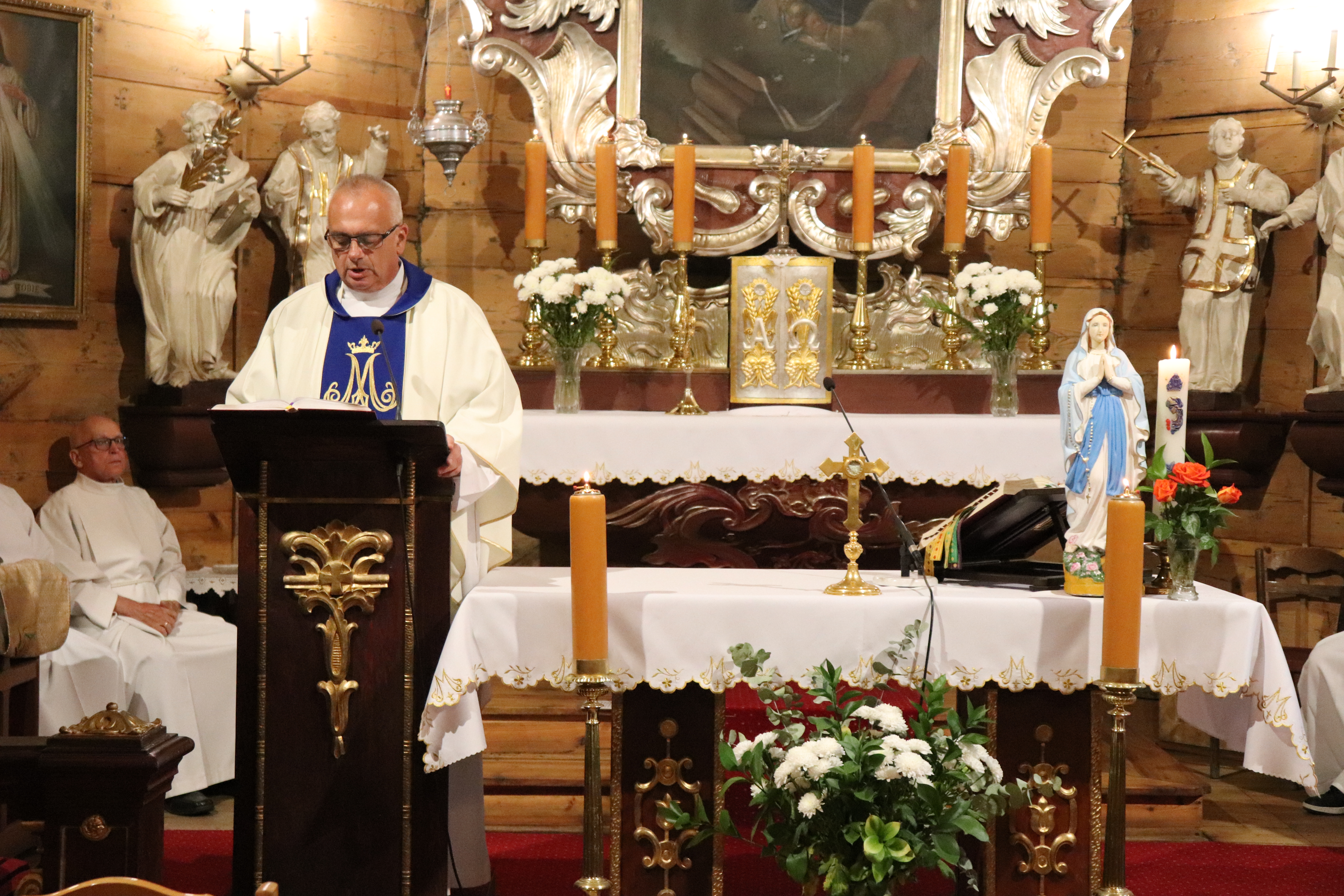 Ołtarz w kościele w Opatówku. Z boku przy drewnianej mównicy stoi ksiądz w biało-niebieskiej szacie, mówi do mikrofonu. W tle figury świętych.