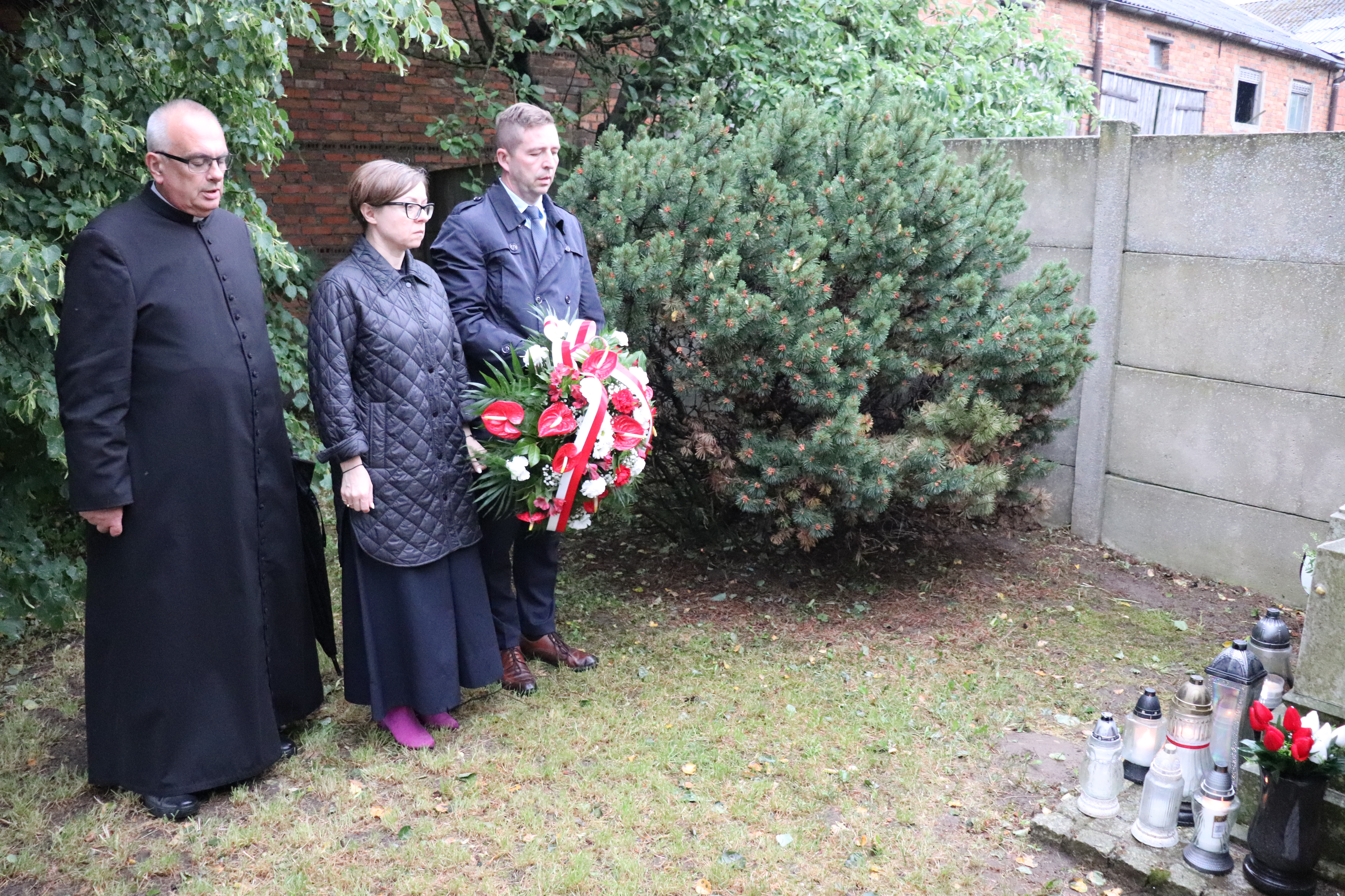 Trzy osoby składają kwiaty przy ścianie z czerwonej cegły, gdzie rozstrzelano Polaków w 1941 r. Od lewej: ksiądz proboszcz, Pani Wiceburmistrz i Pan Burmistrz, trzymający biało-czerwone kwiaty. 